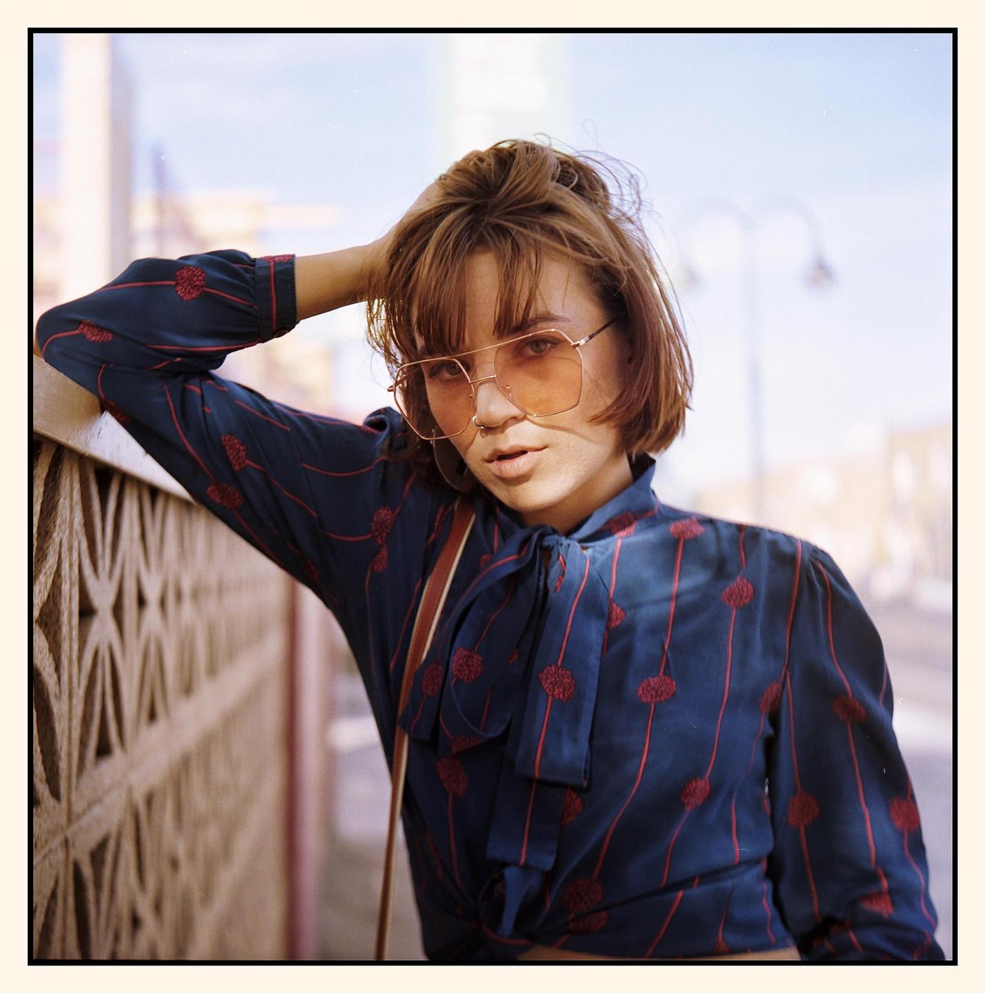 A woman wearing sunglasses and a patterned blouse leans against a wall.