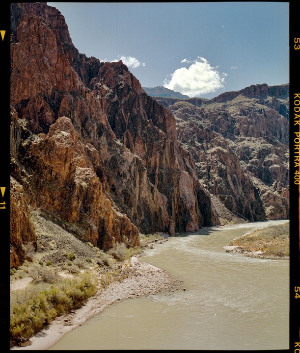 A wide river flows through a canyon with steep, rocky walls under a blue sky with a single cloud.