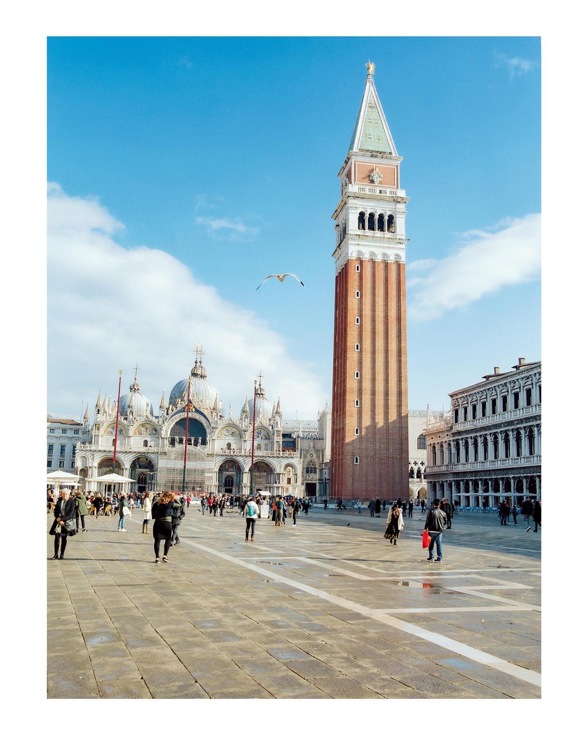 A tall brick bell tower stands over a large square with many people and a large domed building in the background.