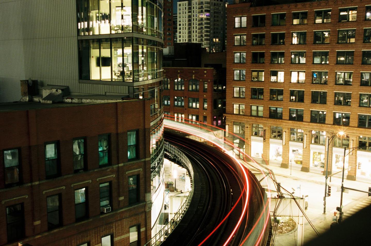 A long exposure captures light trails from a train on an elevated track winding through city buildings at night.