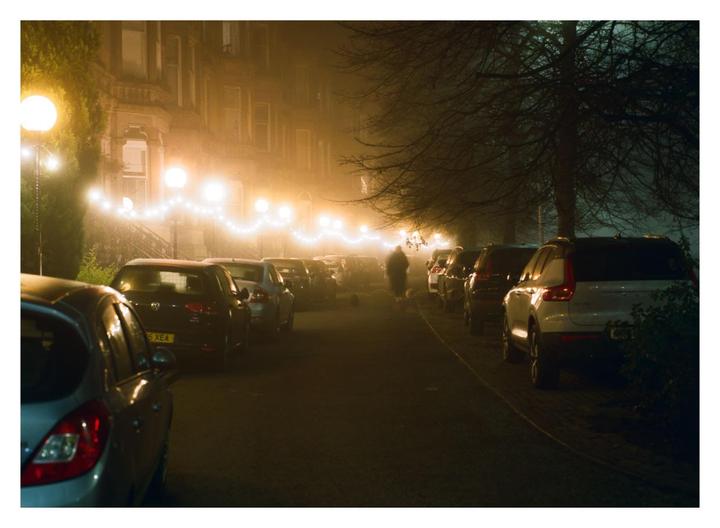A person walks a dog on a foggy street lined with parked cars and illuminated by streetlights and string lights.