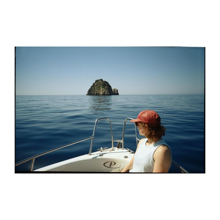 A woman in a red cap sits on a boat looking towards a small rocky island in the distance.