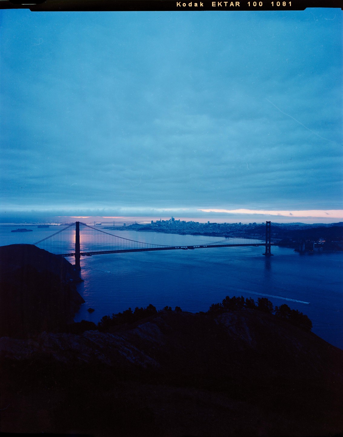 The Golden Gate Bridge spans a body of water with the San Francisco skyline in the distance under a cloudy sky at dawn.