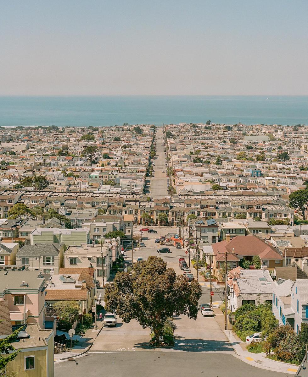 A long street extends through a dense residential area towards the ocean under a clear sky.