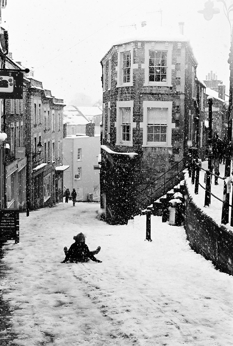 A child sits on a snowy street in a town with stone buildings as snow falls.