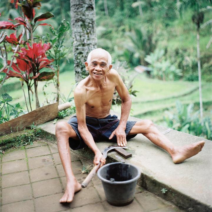 A man sits shirtless on a concrete patio, holding a sickle and smiling at the viewer.