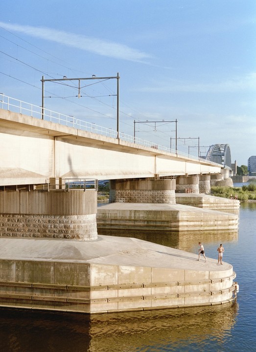 Two men stand on a concrete platform next to a river, with a large bridge overhead and another bridge in the distance.
