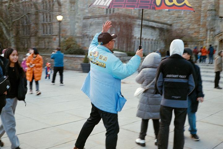 A man in a light blue jacket holds an umbrella and waves to people on a paved street.