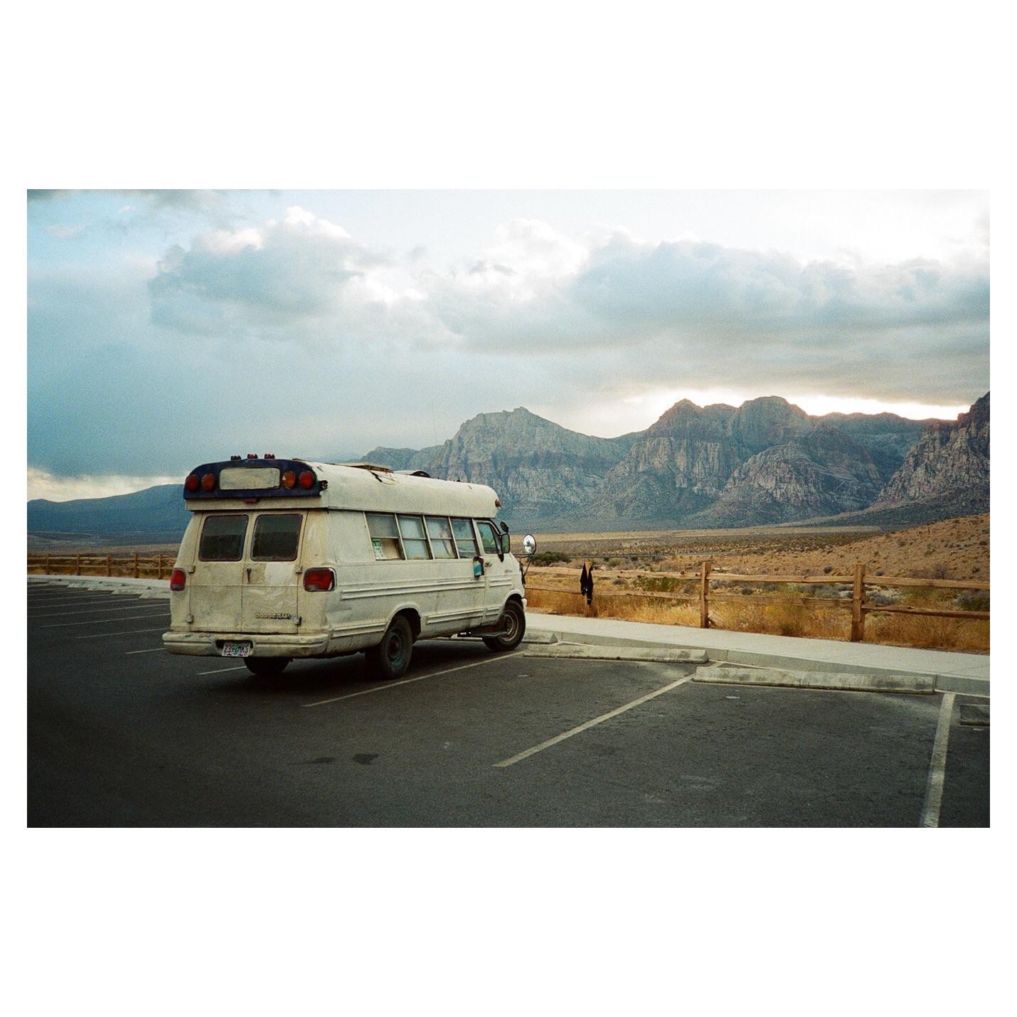 A white van is parked in a parking lot with mountains and a desert landscape in the background under a cloudy sky.