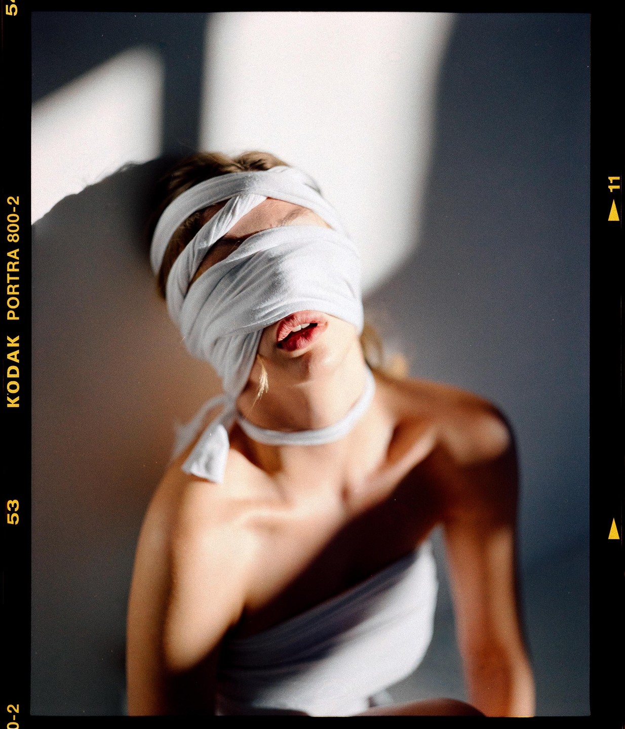 A woman with a white blindfold and white top is bathed in sunlight and shadow.