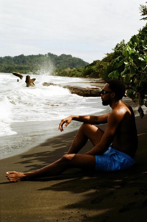 A man in sunglasses and blue swim trunks sits on a dark sand beach with waves crashing in the background.