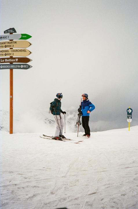 Two skiers stand on a snowy mountain next to a signpost pointing to various ski resorts.