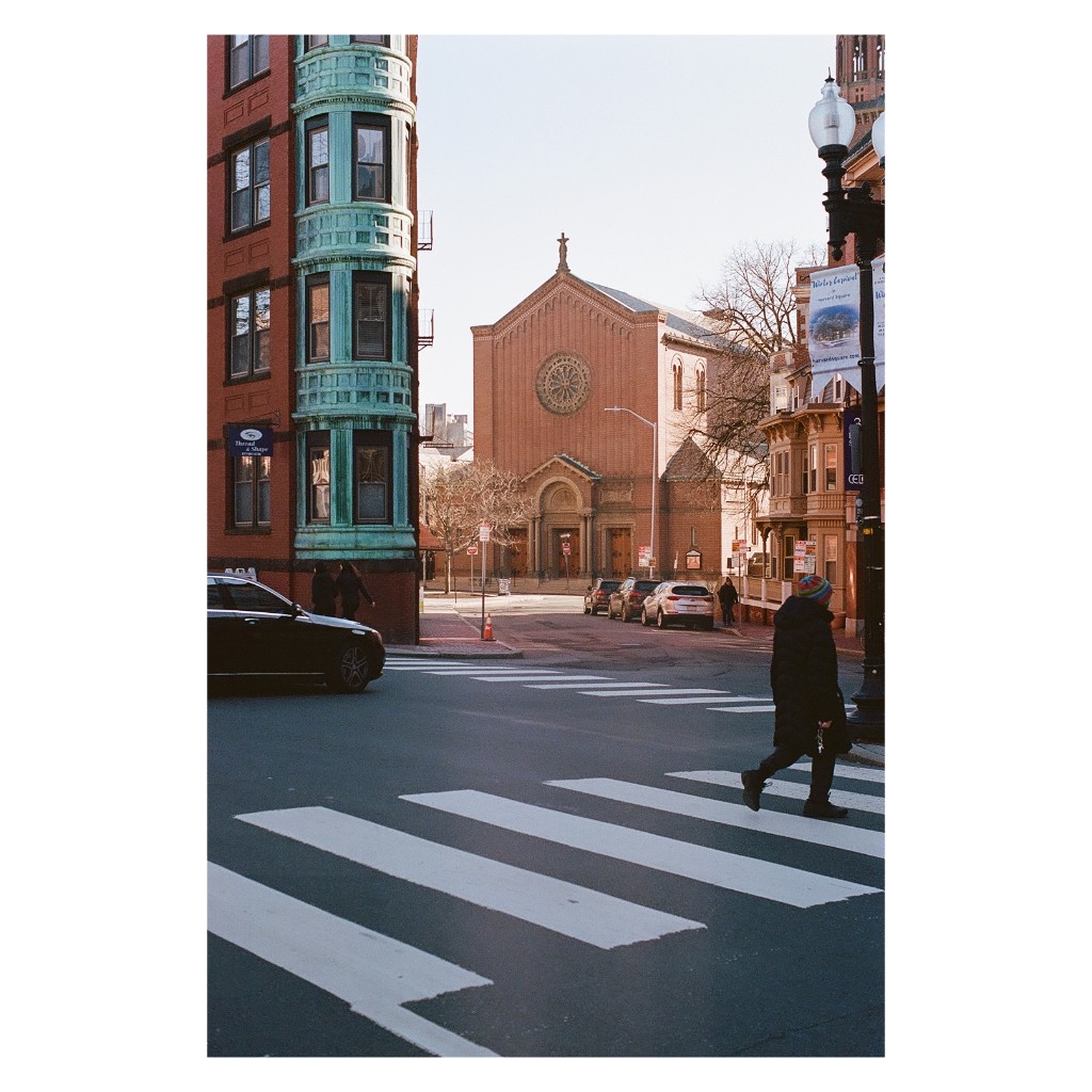 A person crosses a street with a church and buildings in the background.