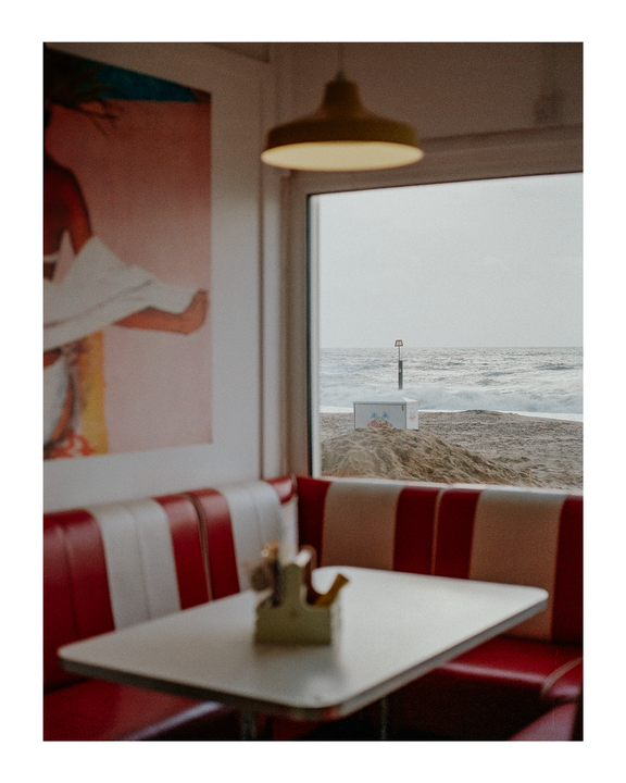A diner booth with red and white striped seating and a table is visible through a window overlooking a beach and ocean.