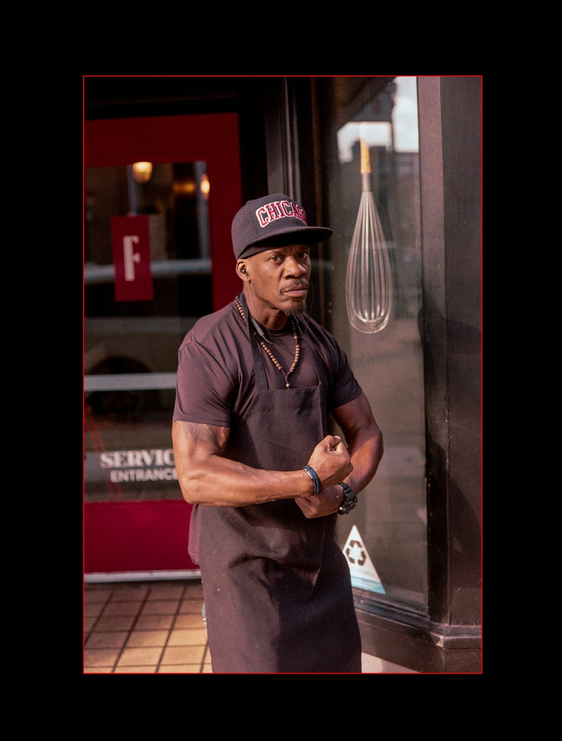 A man wearing an apron and a baseball cap flexes his arm in front of a storefront with a whisk decoration.
