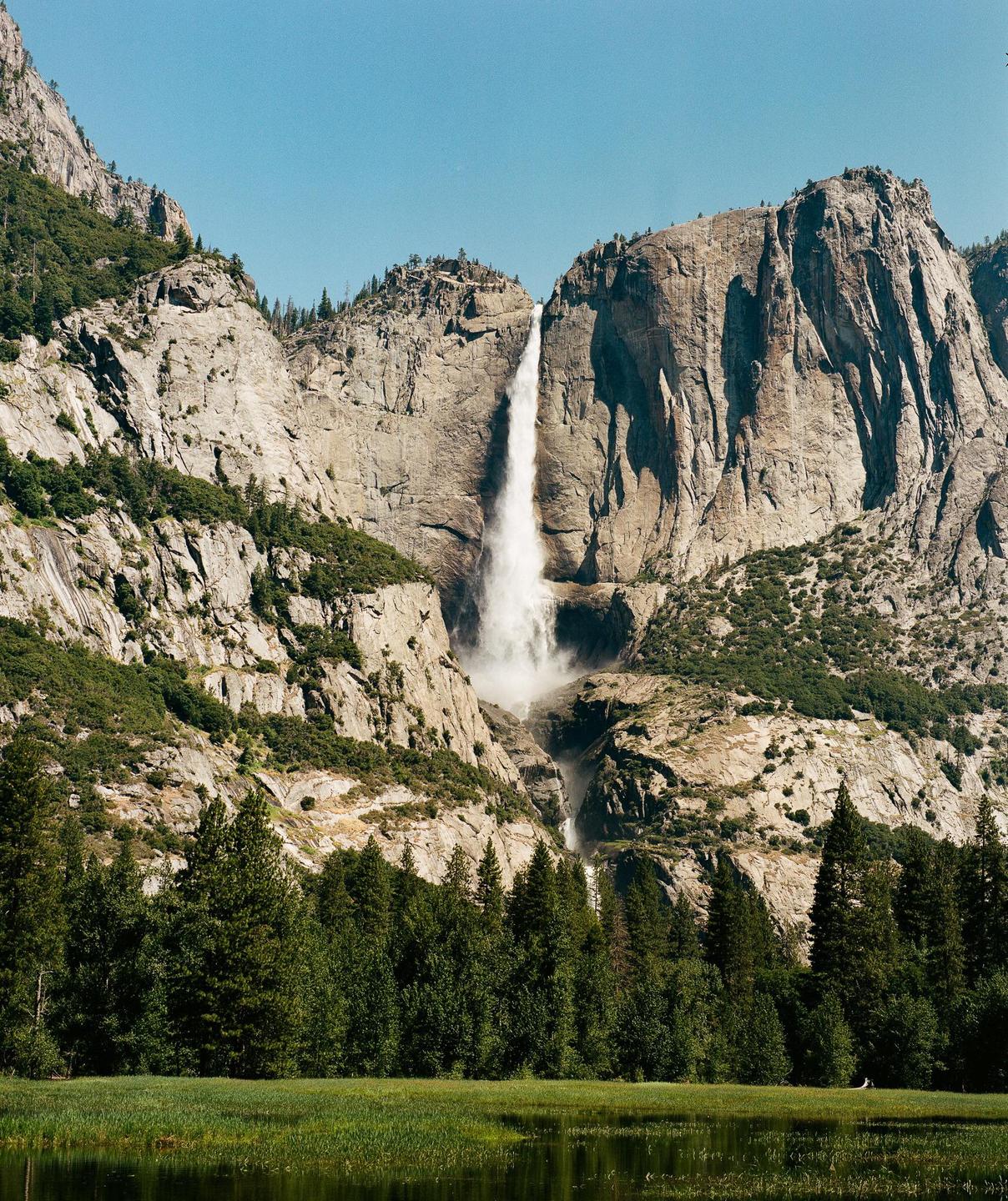 A tall waterfall cascades down a rocky mountain face into a forest and a body of water.