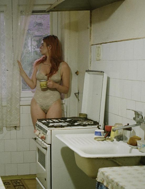 A woman in a bodysuit stands in a kitchen, holding a mug and looking out a window.