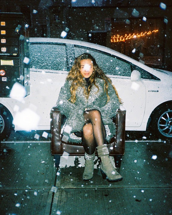 A woman with long, wavy hair sits in a leather armchair on a snowy street at night, with a white car covered in snow behind her.