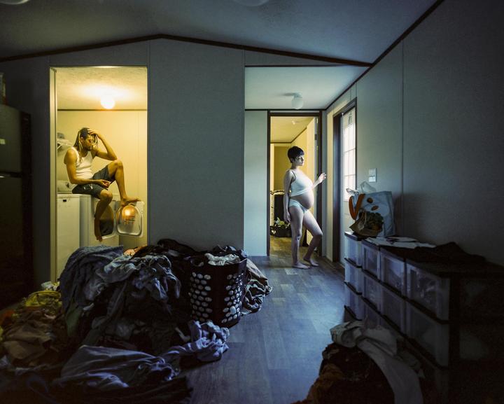 A man sits on a washing machine while a pregnant woman stands in a hallway looking out a door.