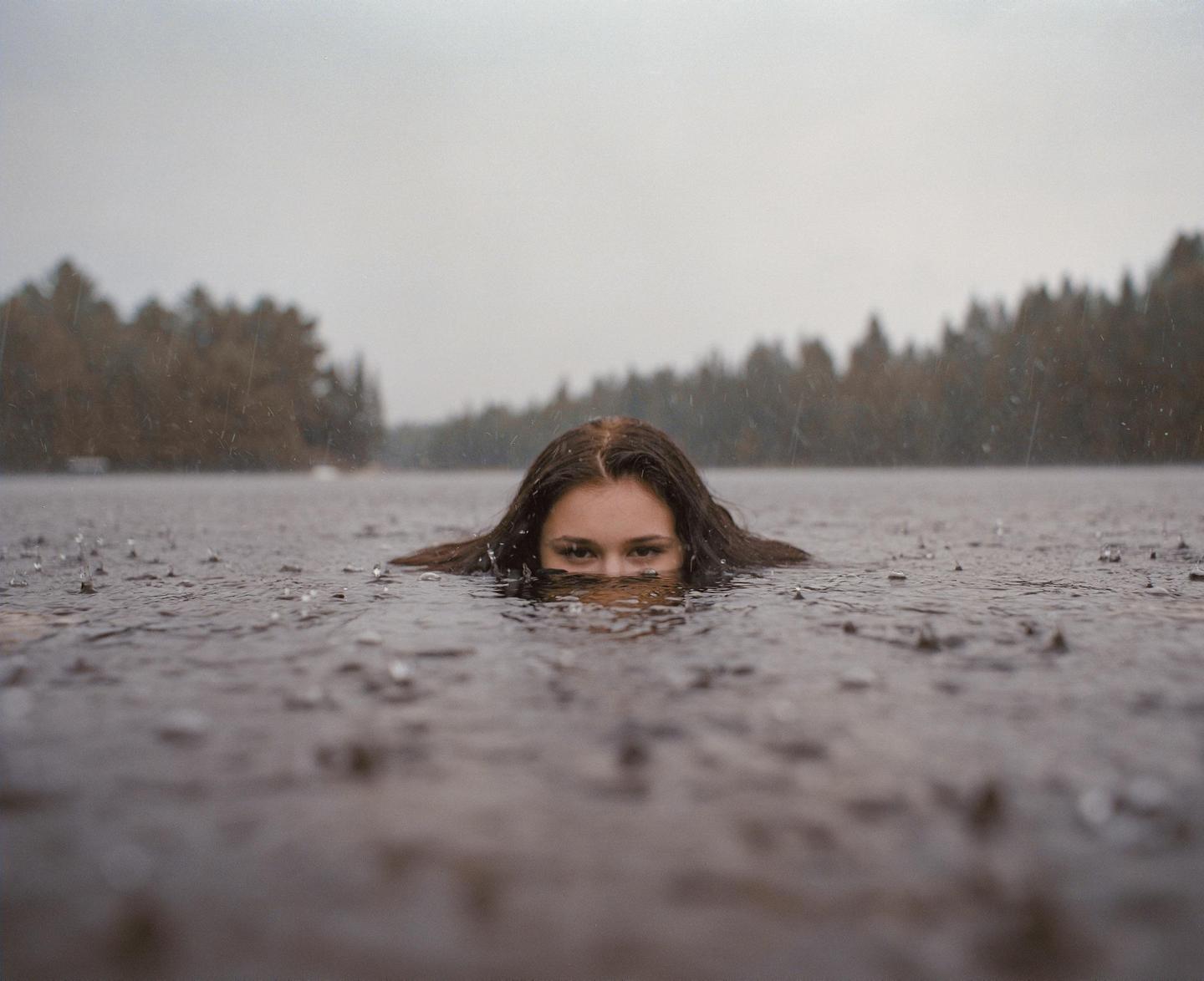 A woman's face is partially submerged in dark water with rain falling around her and a forest in the background.