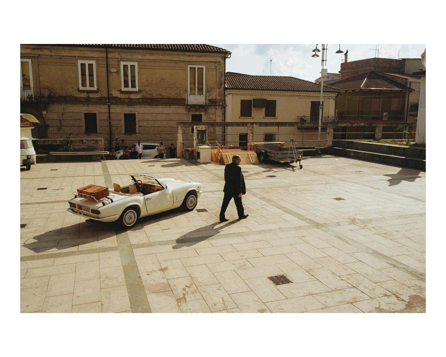 A man walks across a paved town square with a white convertible car parked nearby.