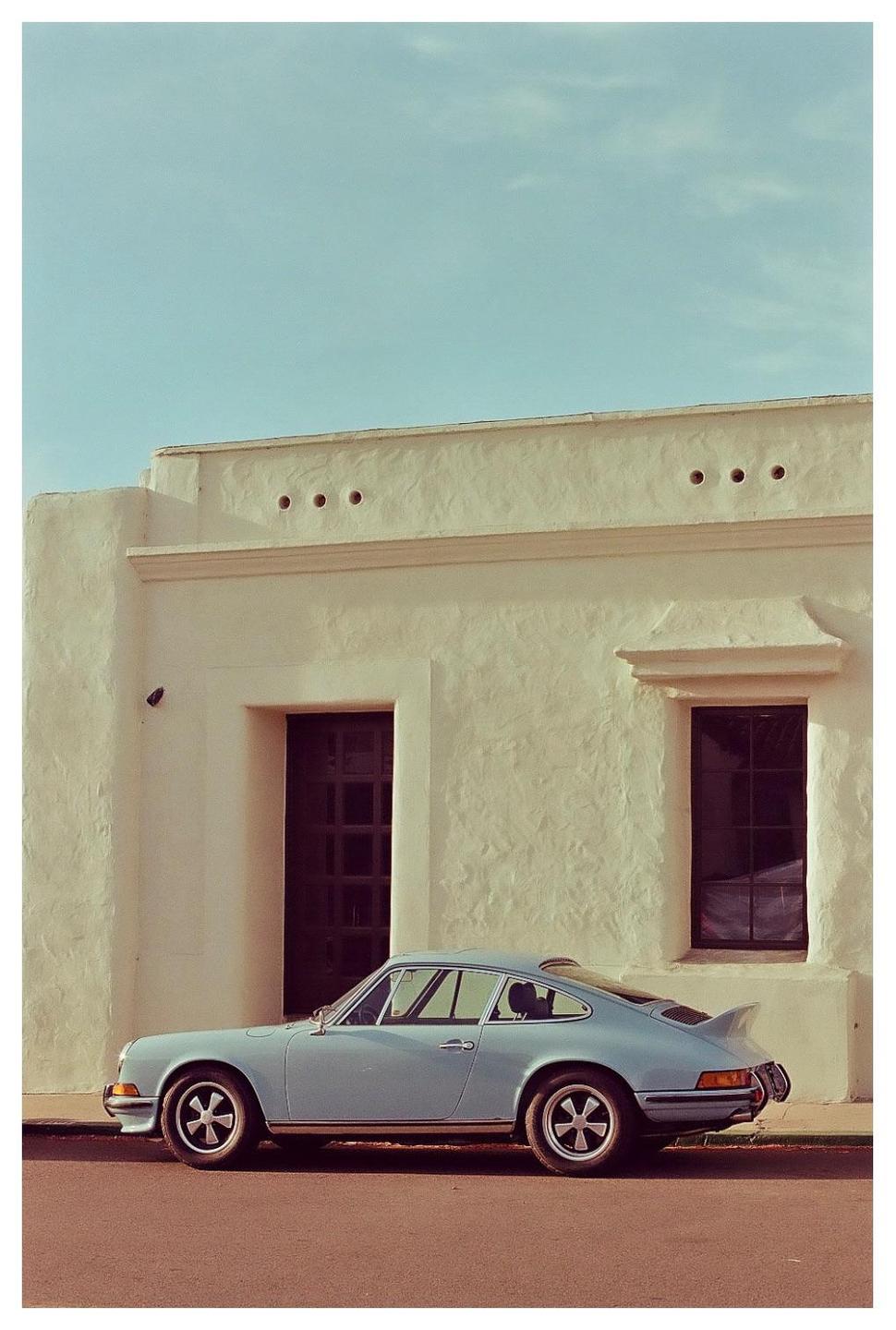 A light blue vintage car is parked on a street next to a cream-colored building with dark windows.