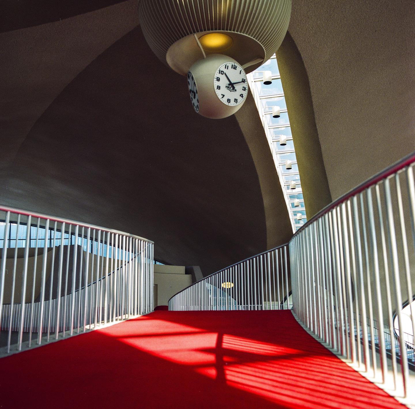 A red carpeted ramp with a metal railing curves under a large clock hanging from a ribbed ceiling.