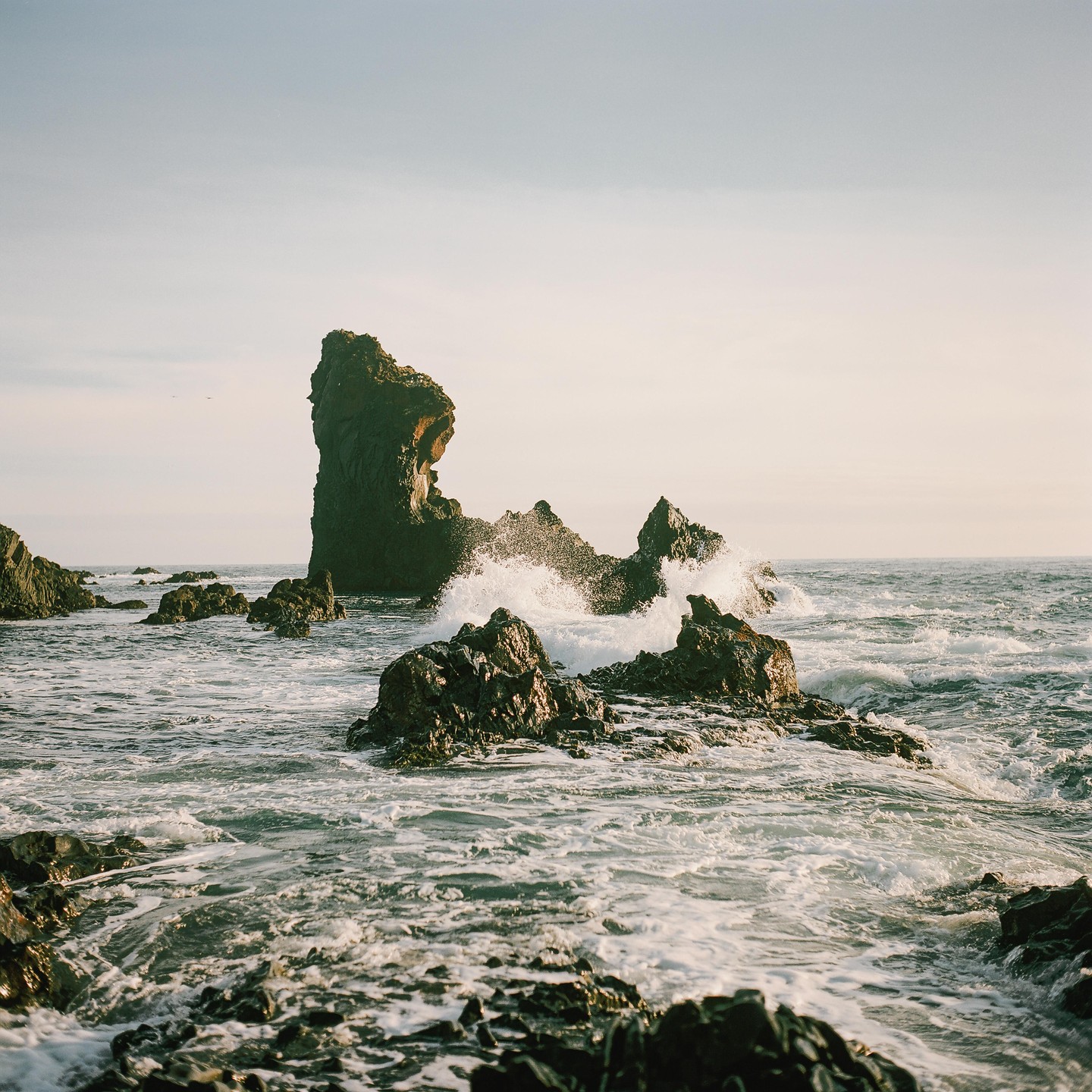 Waves crash against dark, jagged sea stacks and rocks in the ocean under a bright sky.