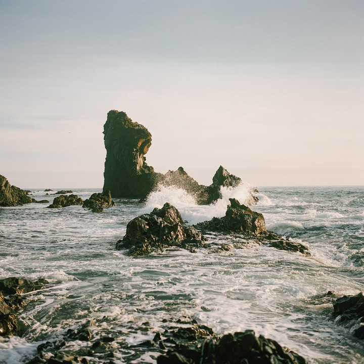 Waves crash against dark, jagged sea stacks and rocks in the ocean under a bright sky.