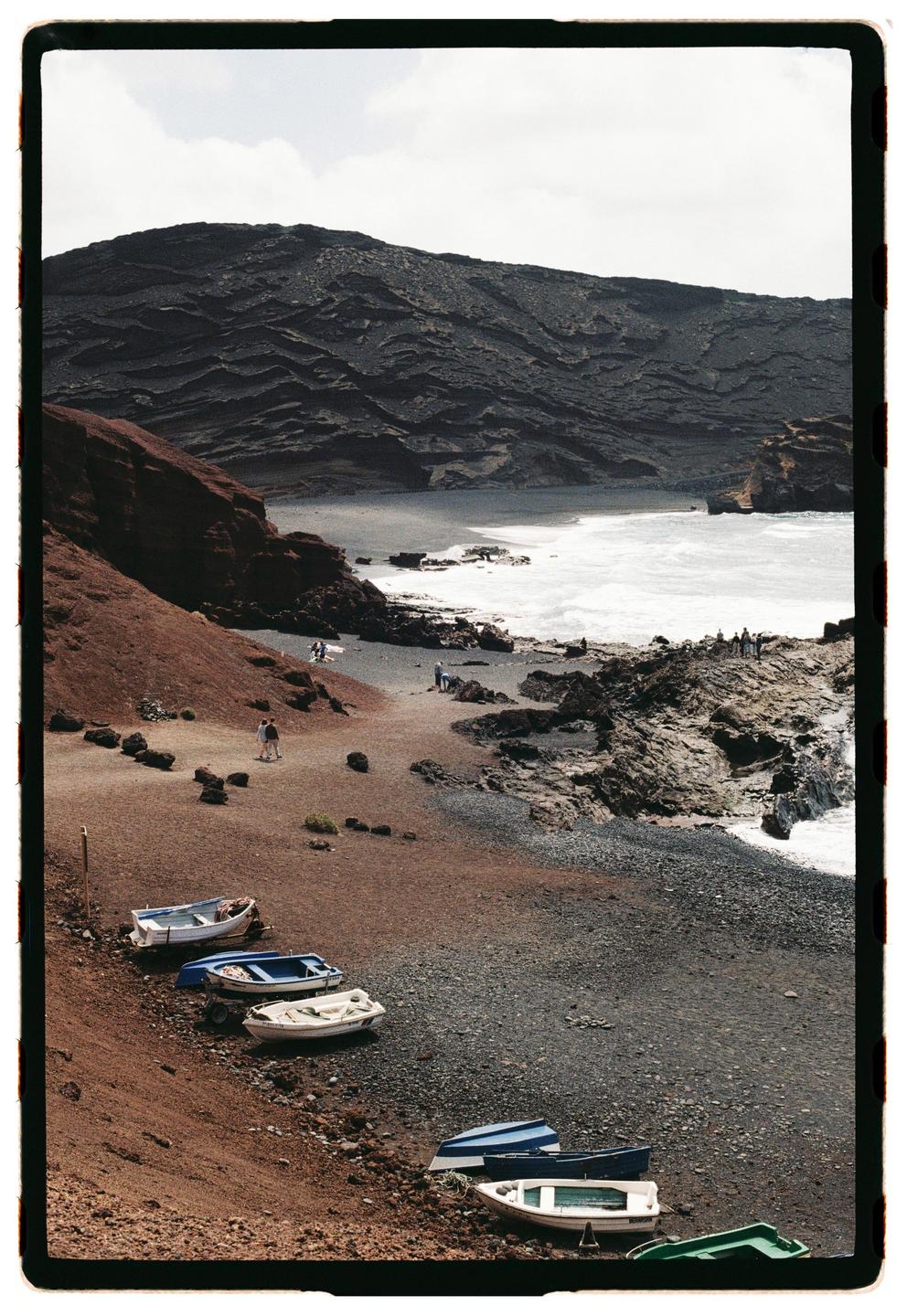 A volcanic beach with small boats on the shore and people walking near the water.