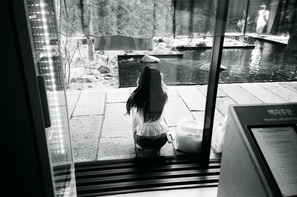 A woman with long hair crouches outside a window next to a body of water.