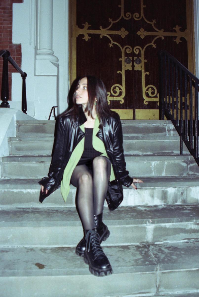 A woman wearing a black leather jacket and black boots sits on stone steps in front of a building with an ornate wooden door.