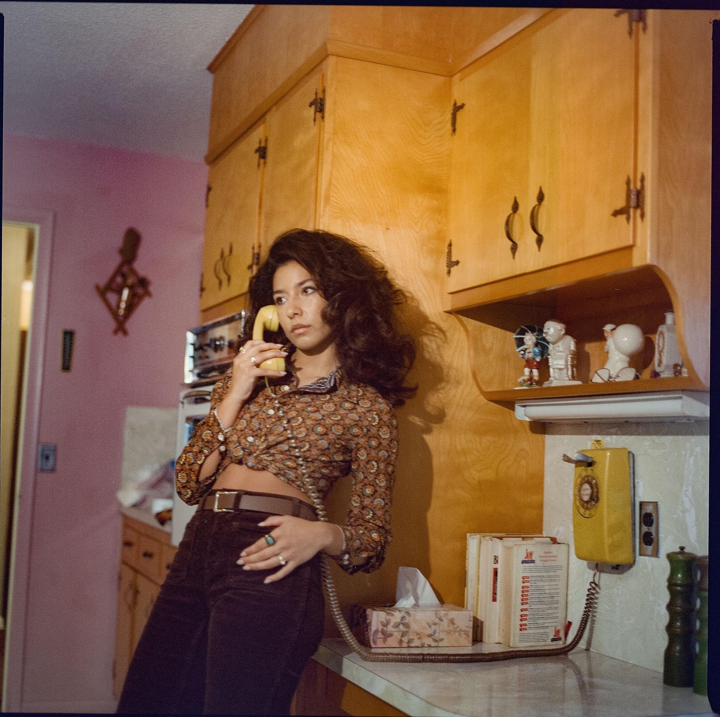 A woman with long, dark hair holds a yellow rotary phone in a kitchen with wooden cabinets.
