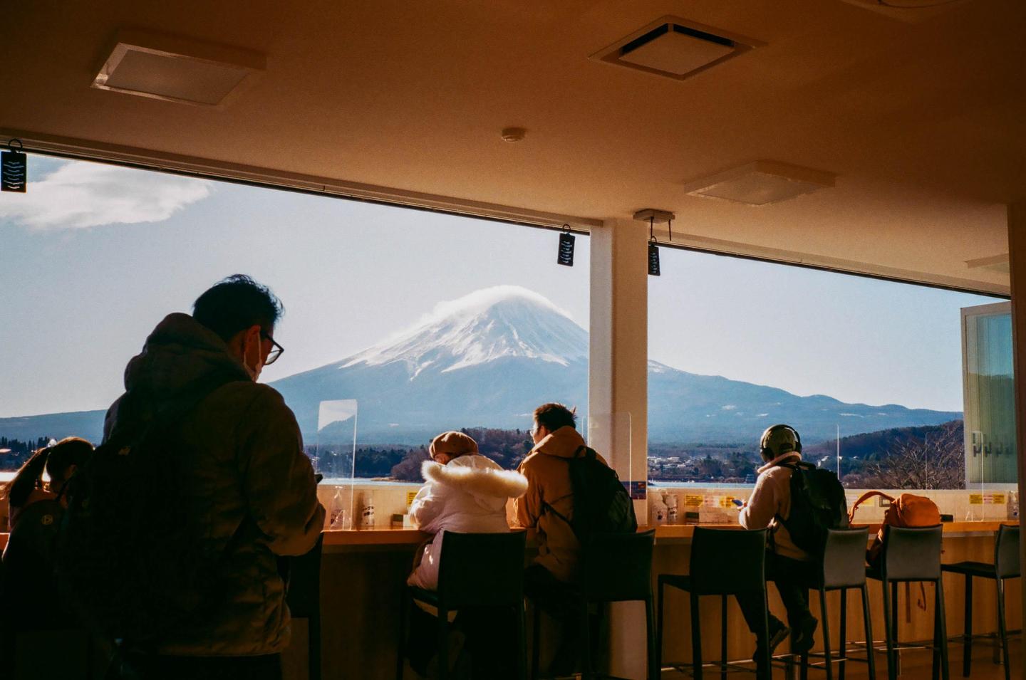People sit at a counter looking out a window at a snow-capped mountain.