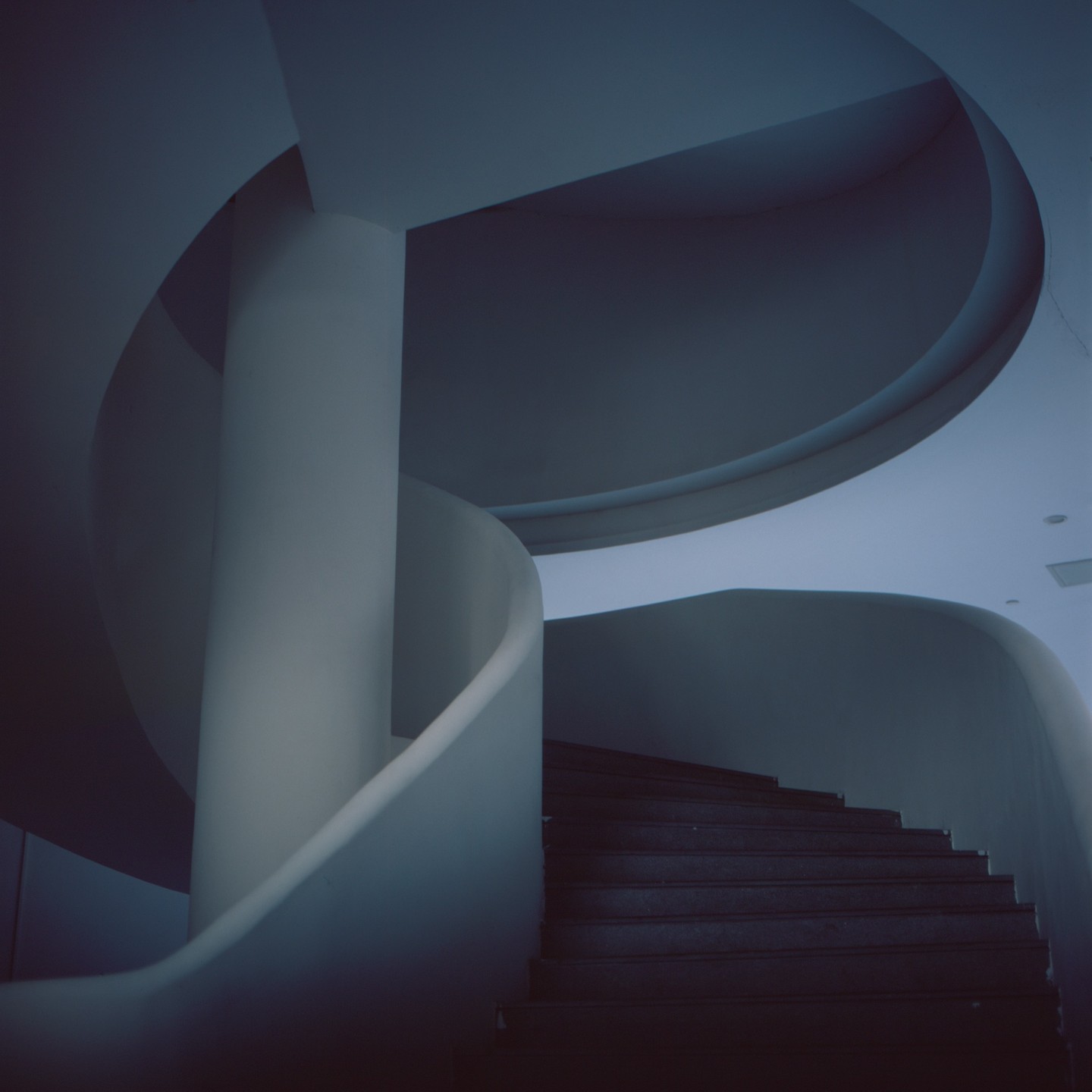 A dark spiral staircase with a central pillar is illuminated by soft light from above.