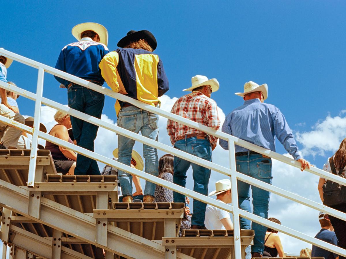 Several men in cowboy hats and jeans stand on bleachers under a bright blue sky with white clouds.