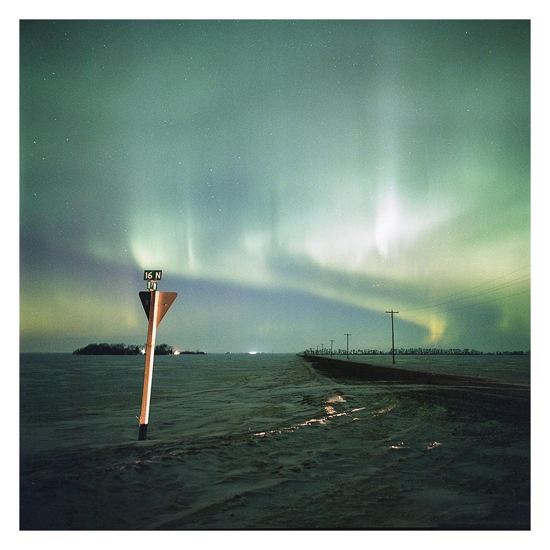The northern lights illuminate a snowy landscape with a road and power lines stretching into the distance.