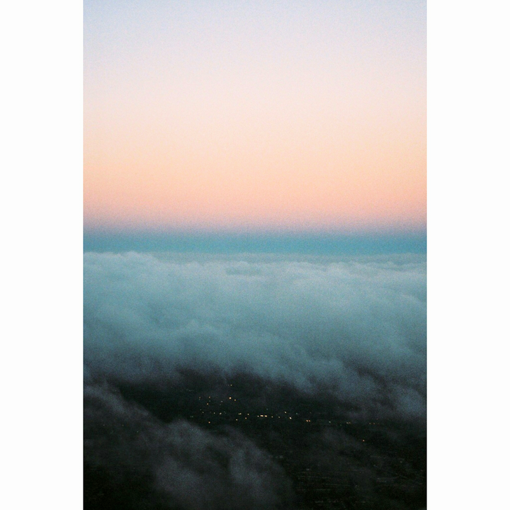A city with lights is visible through a layer of clouds under a colorful sky at dawn.