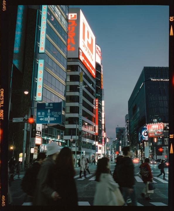 People cross a street in a city at dusk, with neon signs and tall buildings.