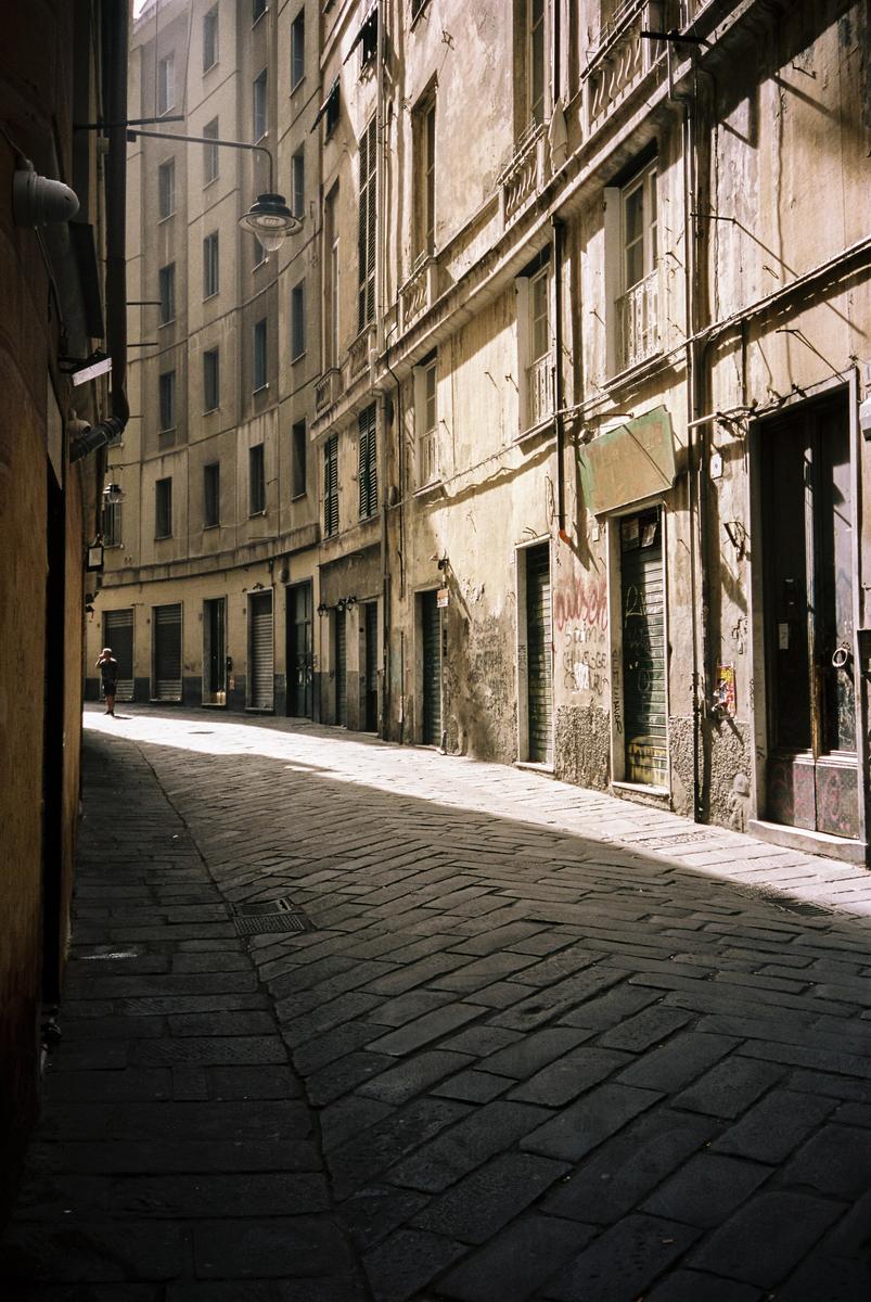 A narrow, curved cobblestone street with tall buildings on either side is illuminated by a diagonal strip of sunlight.
