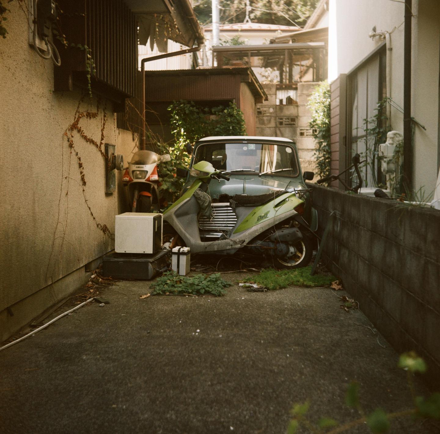 A green scooter and a red motorcycle are parked in a narrow alleyway next to a building.