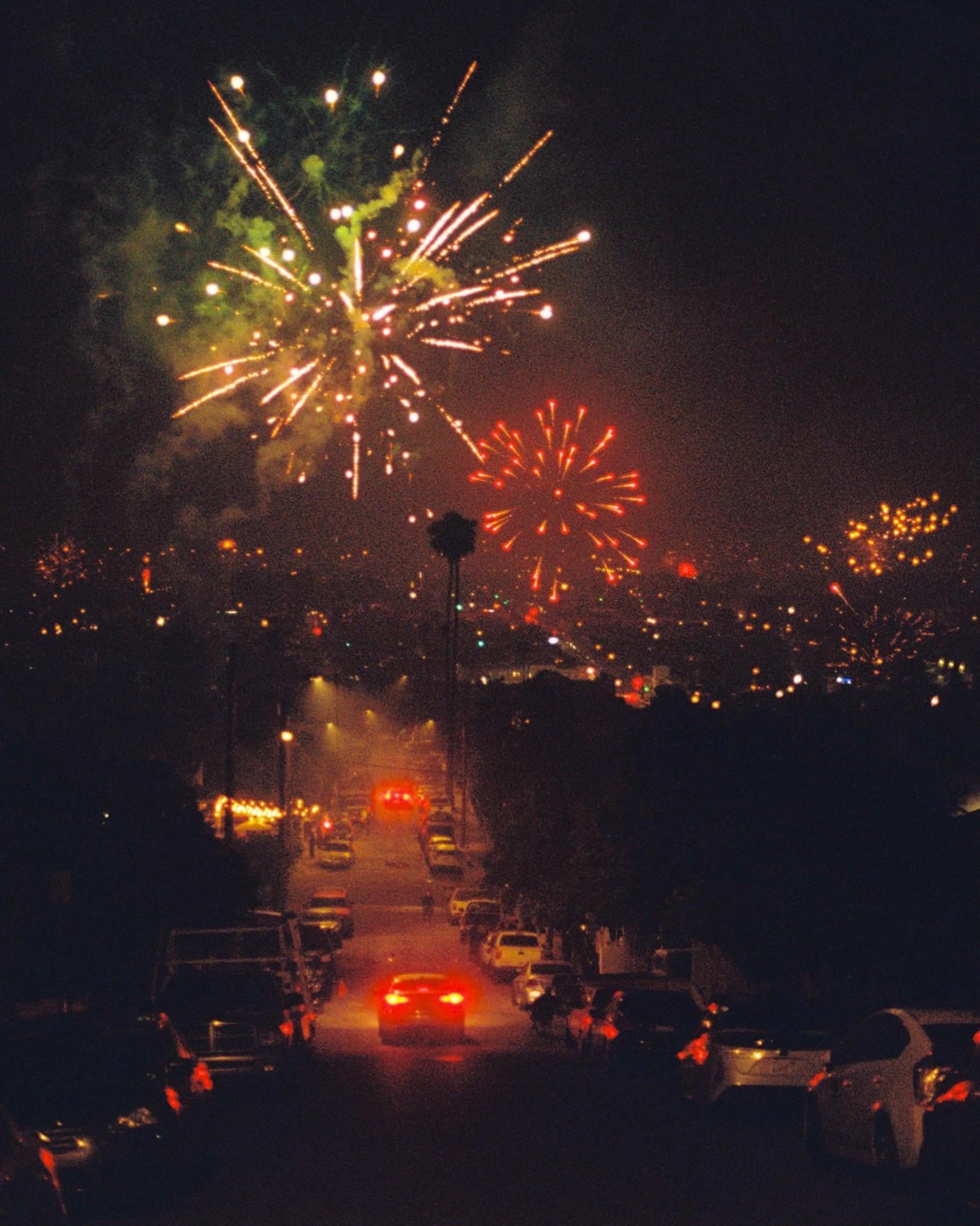 Fireworks explode over a city street at night, with cars parked along the road and a car driving down the street.