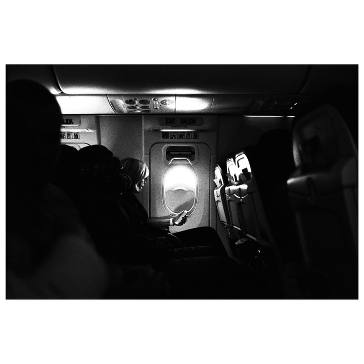 A woman sits in an airplane seat looking at her phone next to a window.