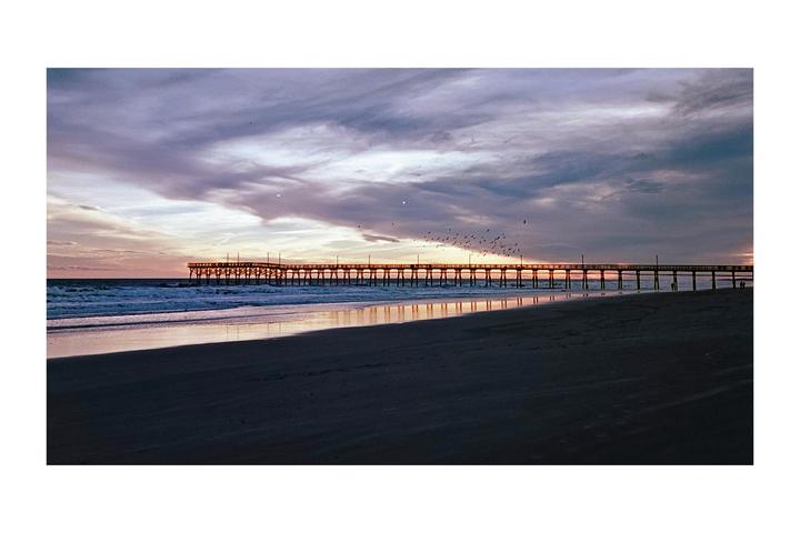 A long pier stretches into the ocean under a dramatic sky at sunset, with birds flying above and waves crashing on the beach.