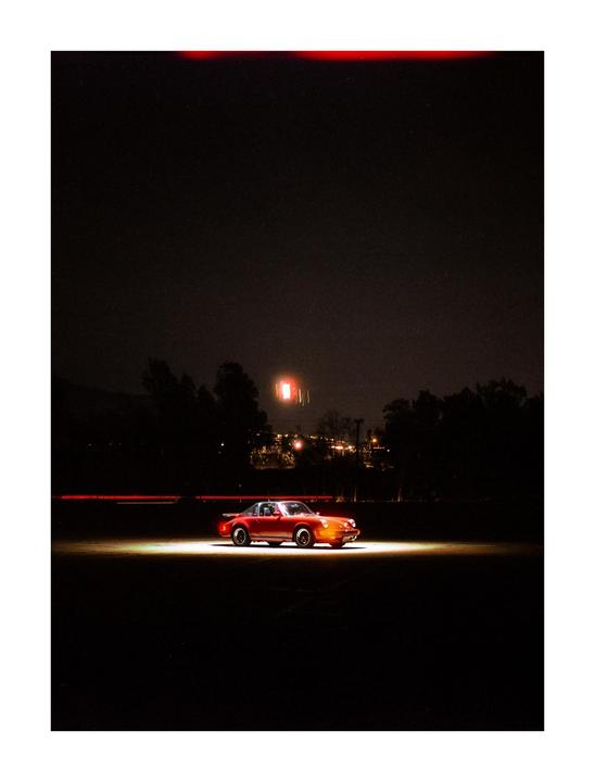A red car is parked on a dark surface with a bright light shining on it from the left.