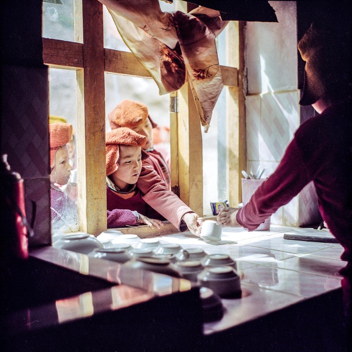 A child monk reaches through a window to receive a cup from another person inside a building.