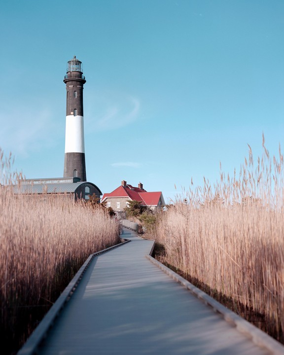 A boardwalk leads through tall grass towards a lighthouse and buildings under a clear sky.