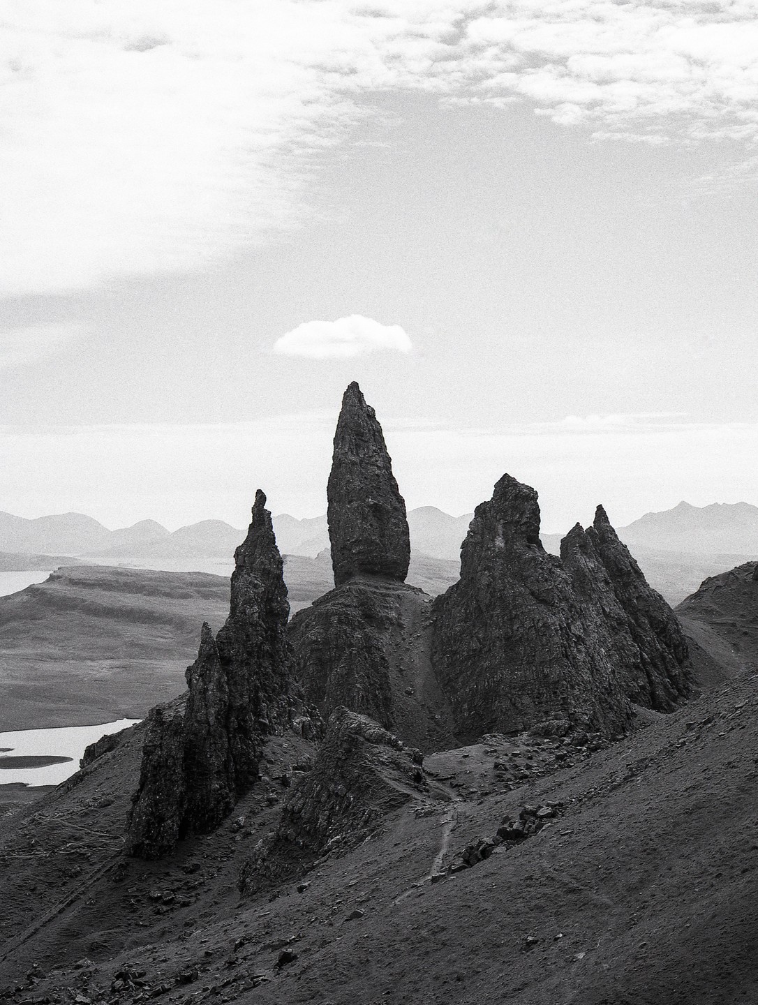A lone cloud floats above a dramatic rock formation on a mountain slope with a body of water in the distance.