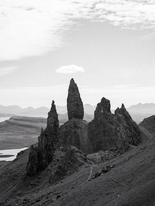 A lone cloud floats above a dramatic rock formation on a mountain slope with a body of water in the distance.