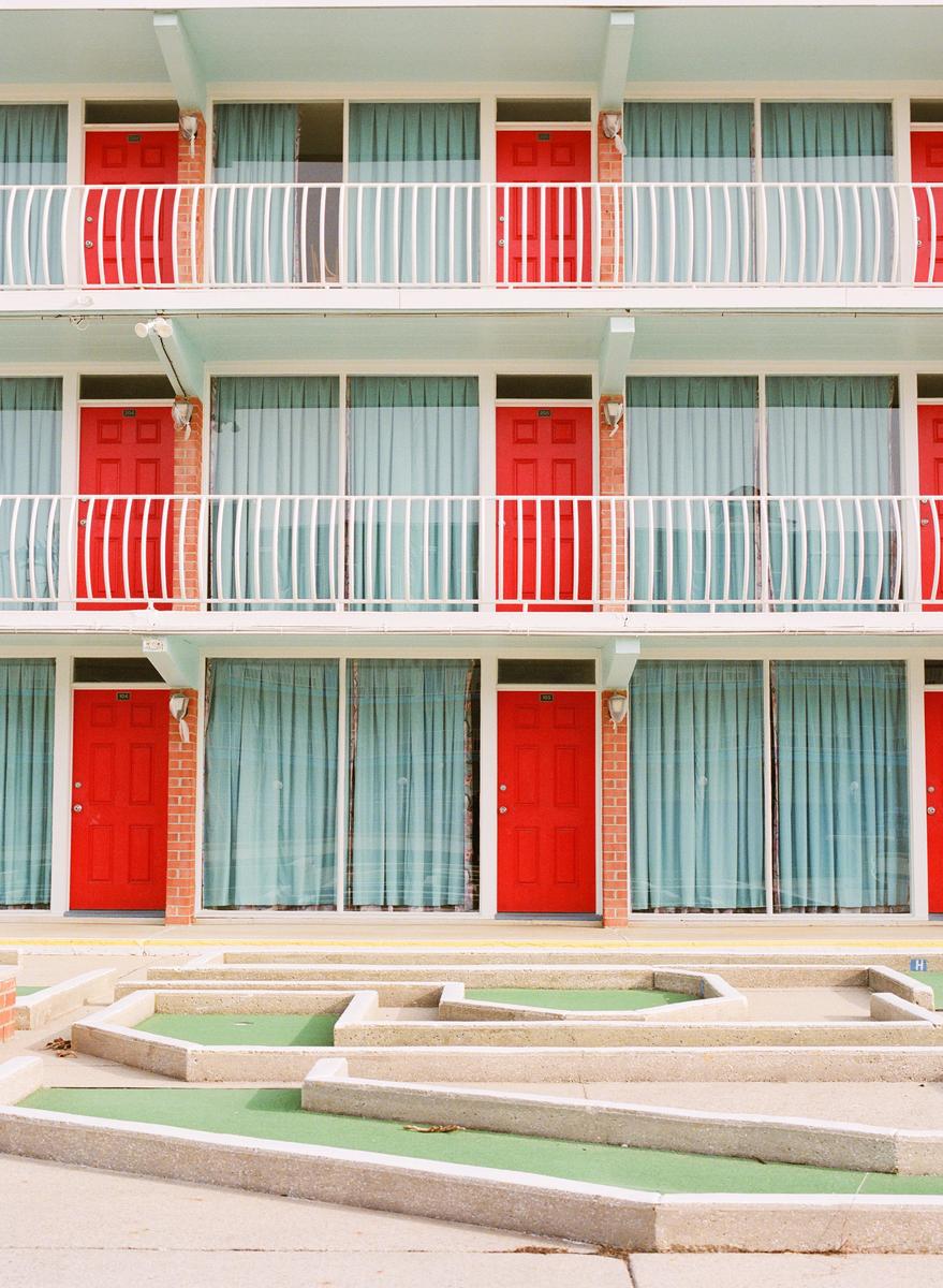 A three-story motel with red doors and light blue curtains has a miniature golf course in front of it.